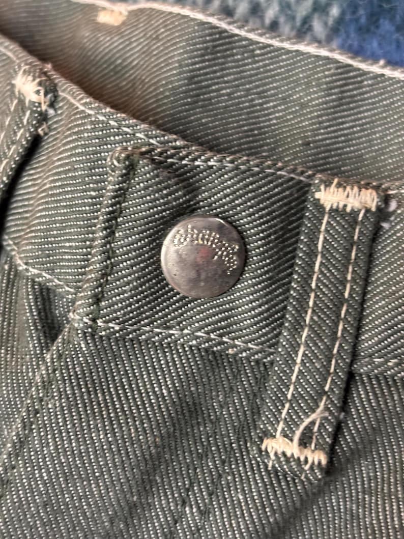 May include: Close-up of denim pants with a silver button and white stitching. The button has an embossed design. The fabric is a dark gray-green color. The stitching is visible along the seams and around the button.