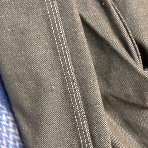 May include: Close-up of a section of dark gray denim fabric with white stitching. The stitching runs vertically, creating a defined seam. The fabric is slightly textured and folded, showing the weave of the material.