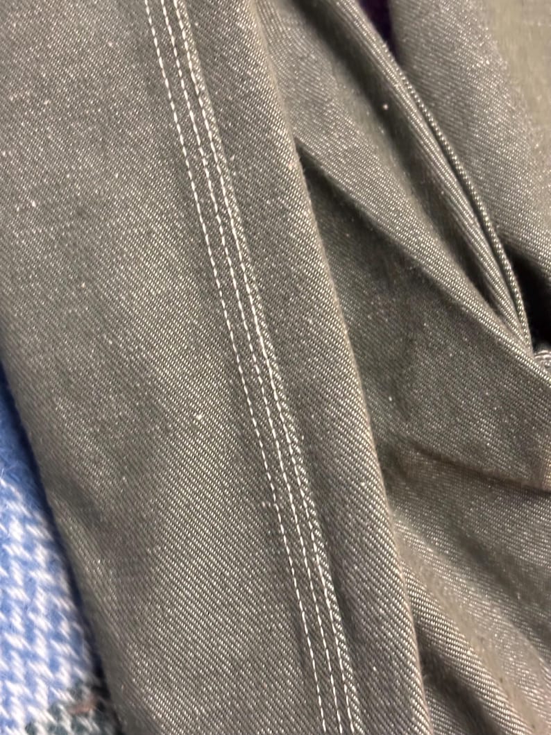 May include: Close-up of a section of dark gray denim fabric with white stitching. The stitching runs vertically, creating a defined seam. The fabric is slightly textured and folded, showing the weave of the material.