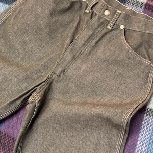 May include: Close-up of brown denim jeans with white stitching. The jeans have a button closure and pockets. The fabric appears to be slightly faded, giving them a vintage look. The jeans are folded.