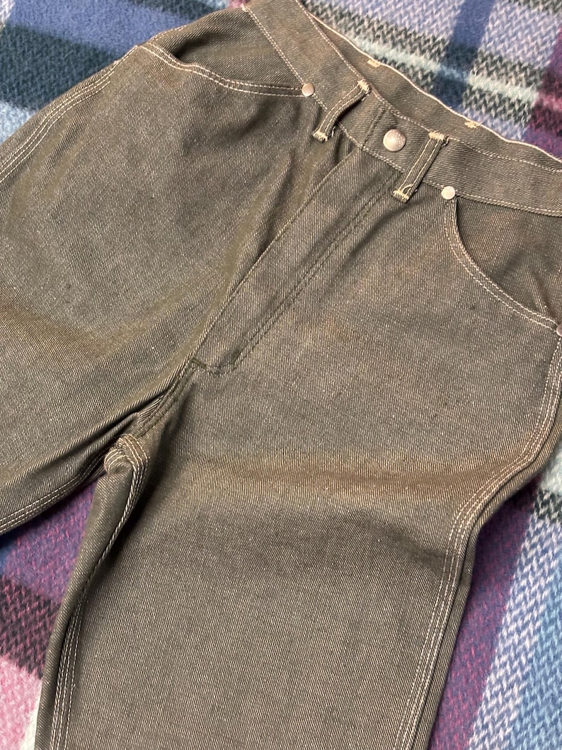 May include: Close-up of brown denim jeans with white stitching. The jeans have a button closure and pockets. The fabric appears to be slightly faded, giving them a vintage look. The jeans are folded.