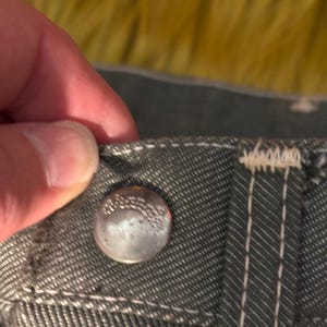 May include: Close-up of a silver button on gray denim fabric. The button is slightly worn and has embossed text. White stitching accents the seams and the buttonhole area. The fabric has a textured appearance.