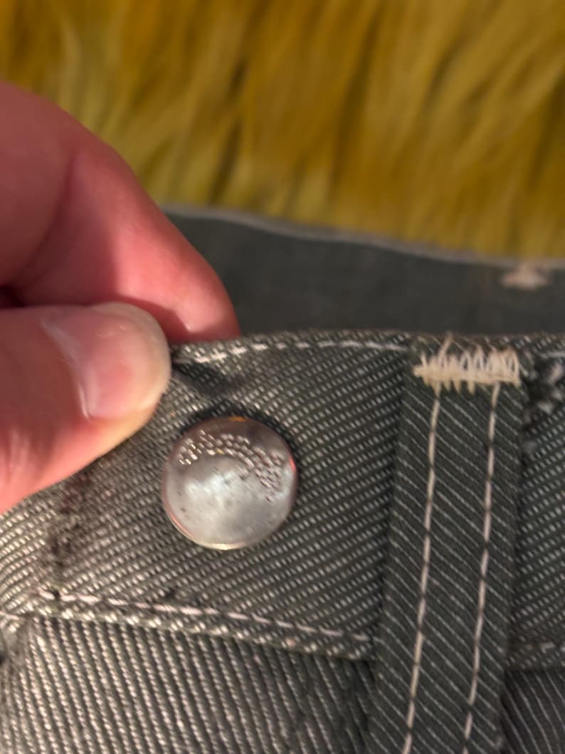 May include: Close-up of a silver button on gray denim fabric. The button is slightly worn and has embossed text. White stitching accents the seams and the buttonhole area. The fabric has a textured appearance.