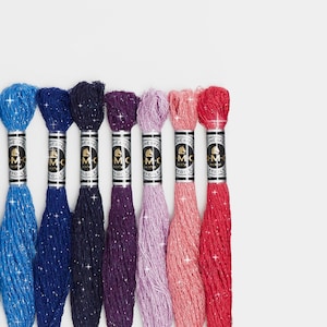 May include: Eight spools of DMC embroidery floss with glitter thread in various colors, including blue, purple, pink, and red. The spools are labeled with the DMC logo.