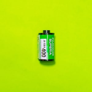Puede incluir: Un rollo de película Fujifilm Superia X-TRA 400, con una etiqueta verde y blanca, y una tapa negra. La película es para 24 copias en color. El fondo es de color verde neón brillante.