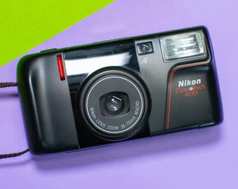 May include: A black Nikon Zoom Touch 400 camera with a 35-70mm zoom lens. The camera has a built-in flash and a red accent on the side.