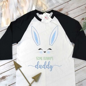 May include: White and black baseball tee shirt with a blue bunny face and the text "Some bunny's daddy" in green. Two gold arrows are crossed in front of the shirt.
