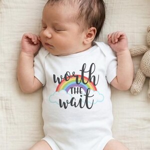 May include: A white short-sleeved baby bodysuit featuring a graphic of a rainbow and clouds with the black text "worth THE wait" centered on the chest. The bodysuit is worn by a sleeping infant lying in a woven wicker basket with a small knit teddy bear.