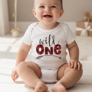 May include: A white short-sleeved baby bodysuit featuring the text "wild one" in black script and red and black buffalo plaid block lettering, with a small arrow graphic underneath the word "one" printed on the front of the garment.