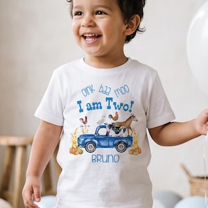 May include: A white short-sleeved t-shirt featuring a blue truck graphic with farm animals, pumpkins, and the text "oink baa moo I am Two!" and "BRUNO" in blue and brown lettering. The shirt is paired with light beige linen-style trousers.