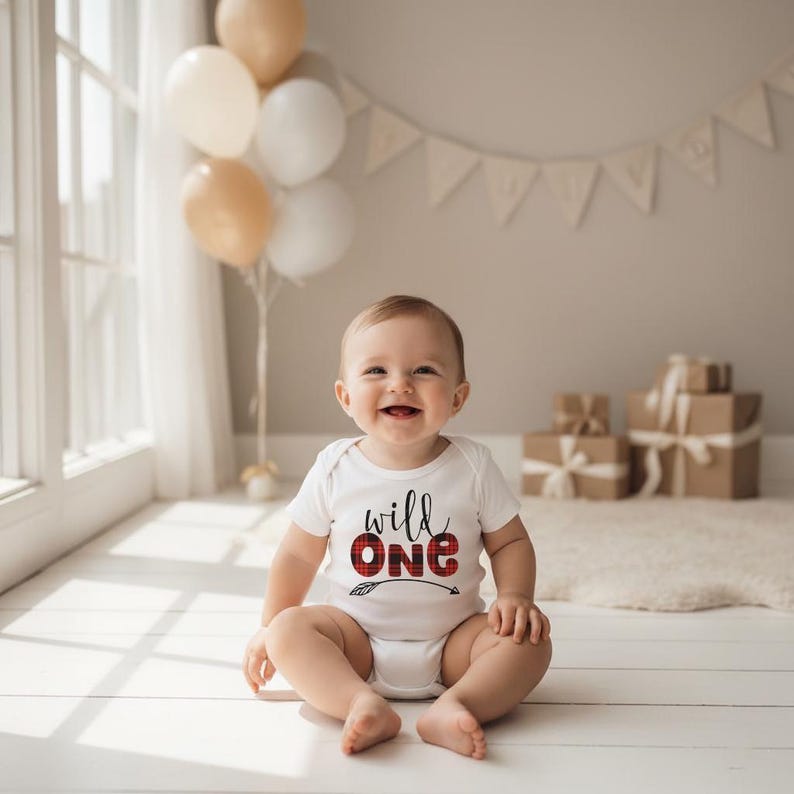 May include: A white short-sleeved baby bodysuit featuring the text "wild one" in black script and red and black buffalo plaid block lettering, with a small arrow graphic underneath the word "one" printed on the front of the garment.