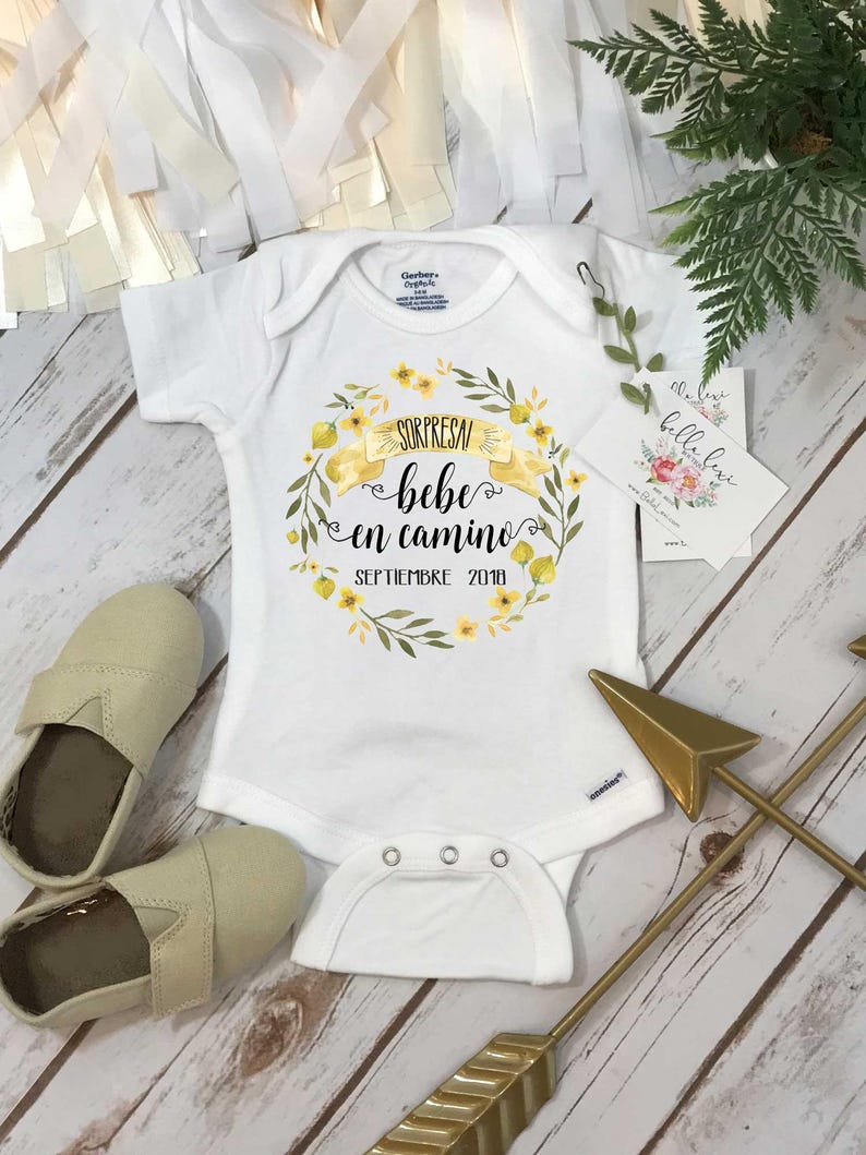 Puede incluir: Un body blanco para beb&eacute; con una corona de flores amarillas y el texto "Sorpresa bebe en camino Septiembre 2019".