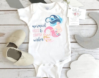 Peut inclure: Un body pour bébé blanc à manches courtes orné d'une sirène aux cheveux bleus et à la queue rose, accompagné du texte "mermaid KISSES & STARFISH wishes" en lettres bleues et roses, accompagné d'une paire de chaussures beiges à enfiler.