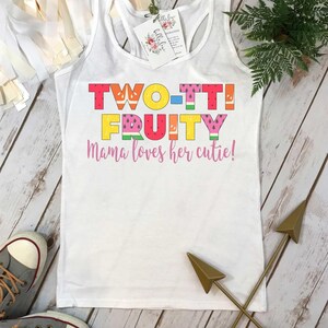 May include: A white racerback tank top featuring the colorful text TWO-TTI FRUITY and the phrase Mama loves her cutie! in pink script. The letters are decorated with various fruit patterns including watermelon, orange, and kiwi designs.