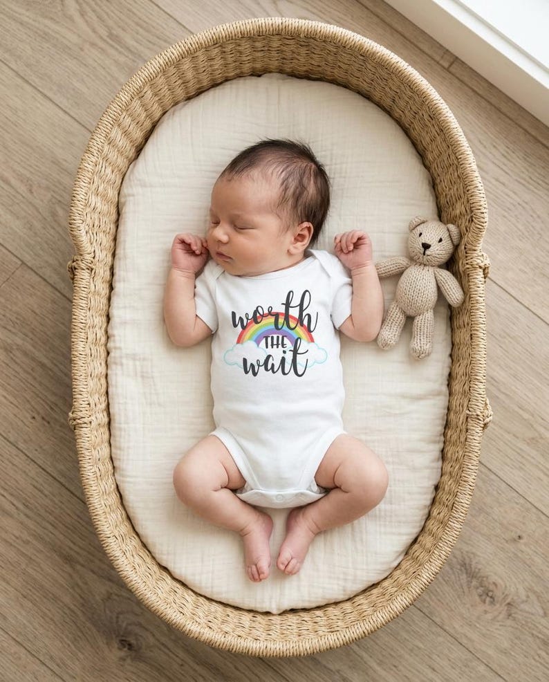 May include: A white short-sleeved baby bodysuit featuring a graphic of a rainbow and clouds with the black text "worth THE wait" centered on the chest. The bodysuit is worn by a sleeping infant lying in a woven wicker basket with a small knit teddy bear.