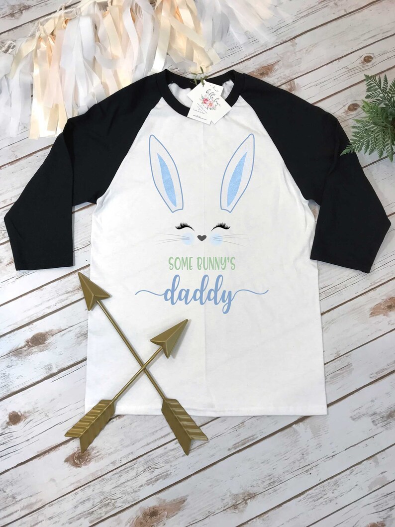 May include: White and black baseball tee shirt with a blue bunny face and the text "Some bunny's daddy" in green. Two gold arrows are crossed in front of the shirt.