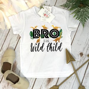 May include: A white short-sleeved t-shirt featuring a graphic of yellow dinosaurs and green tropical leaves. The text reads BRO of the Wild Child in black lettering. A small white brand tag is attached to the neckline of the garment.