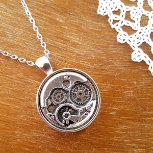 Può includere: Una collana con ciondolo circolare color argento che mostra un meccanismo di orologio a vista con ingranaggi e ruote dentate metalliche. Il ciondolo è appeso a una sottile catena a maglie color argento.