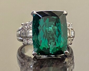 May include: A silver ring featuring a large, emerald-cut, deep green gemstone. The ring has smaller, clear gemstones on either side of the main stone. The ring is set on a reflective surface.