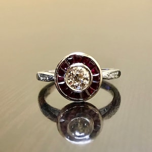 May include: A silver ring featuring a round, bezel-set diamond encircled by a halo of red rubies.