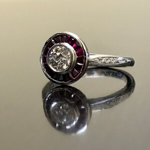 May include: A silver ring with a round center stone surrounded by a halo of red rubies. The band is set with smaller diamonds.