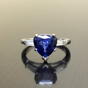 May include: A heart-shaped blue sapphire engagement ring set in a silver band with two smaller diamonds on either side of the sapphire.