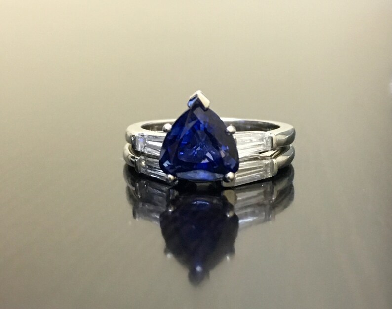 May include: A silver ring set with a large, dark blue, triangular sapphire gemstone. The sapphire is surrounded by smaller, clear, baguette-cut diamonds.