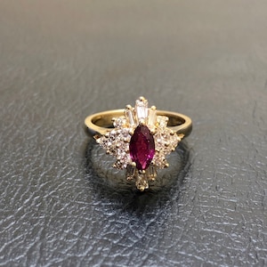 Op de afbeelding: Een gouden ring met een grote, donkerpaarse edelsteen omringd door kleinere, heldere edelstenen.