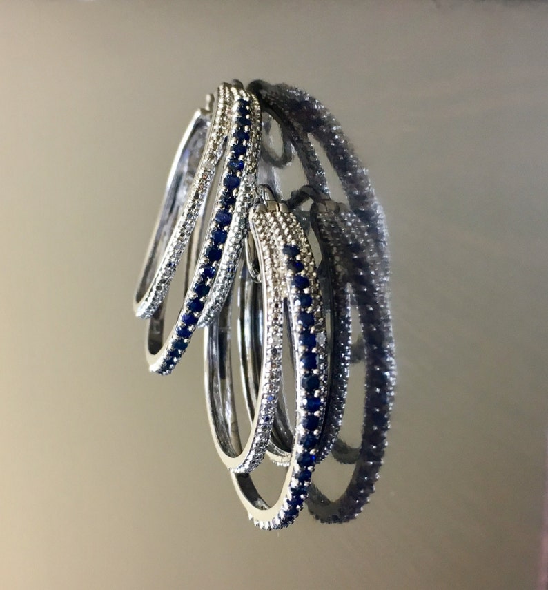 May include: Four silver hoop earrings with blue gemstones set in the metal. The earrings are stacked on top of each other.