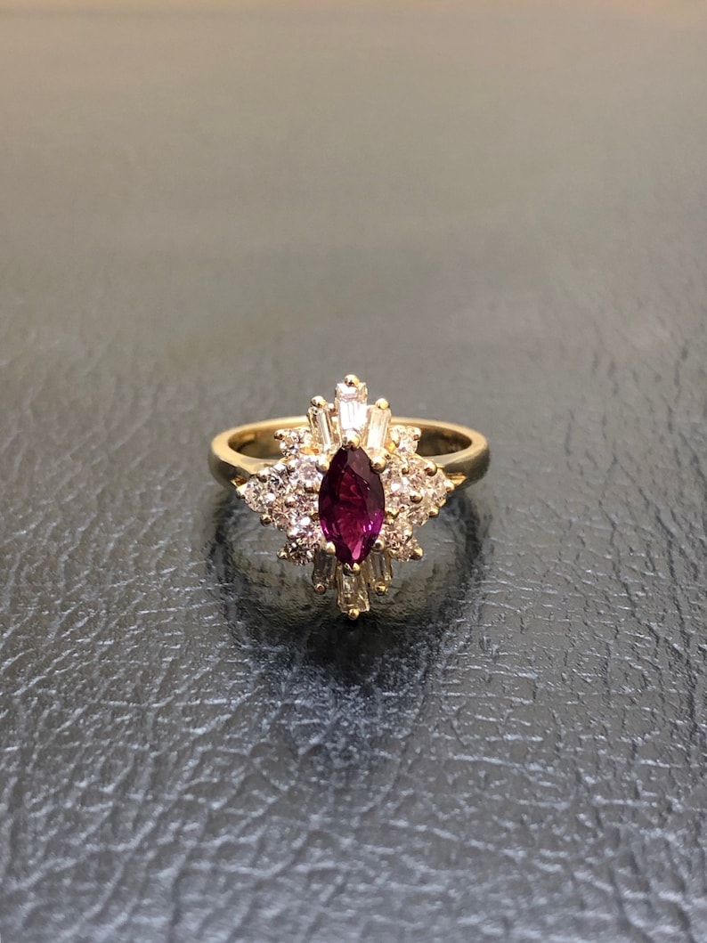 Op de afbeelding: Een gouden ring met een grote, marquise-vormige robijn omringd door kleinere diamanten. De ring heeft een delicaat, bloemig ontwerp.