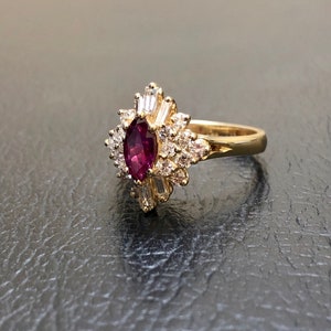 May include: A gold ring featuring a large, oval ruby surrounded by smaller diamonds. The ring has a delicate, floral design.