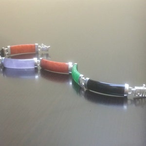 May include: A silver bracelet with four different colored jade stones. The stones are green, black, purple, and orange. The bracelet has a silver clasp.