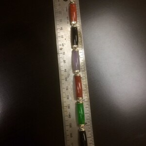 May include: A bracelet with five gemstone beads in red, black, purple, red, and green. The beads are strung on a silver chain and measured against a ruler.