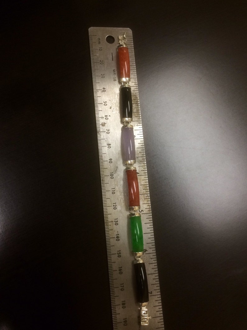 May include: A bracelet with five gemstone beads in red, black, purple, red, and green. The beads are strung on a silver chain and measured against a ruler.