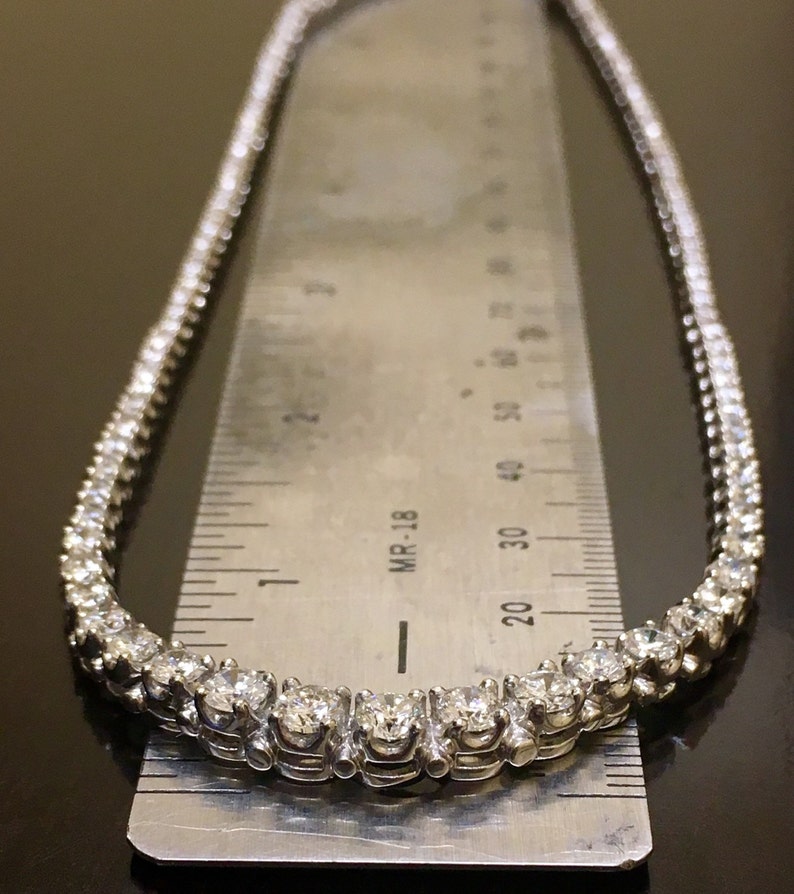 May include: A silver tennis necklace with a row of small, round diamonds. The necklace is laid on a ruler, showing the length to be about 18 inches.