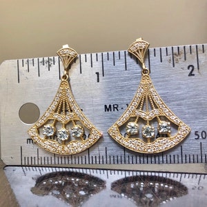 May include: A pair of gold earrings with a fan-shaped design and clear stones. The earrings are about 1 inch long and are shown next to a ruler.