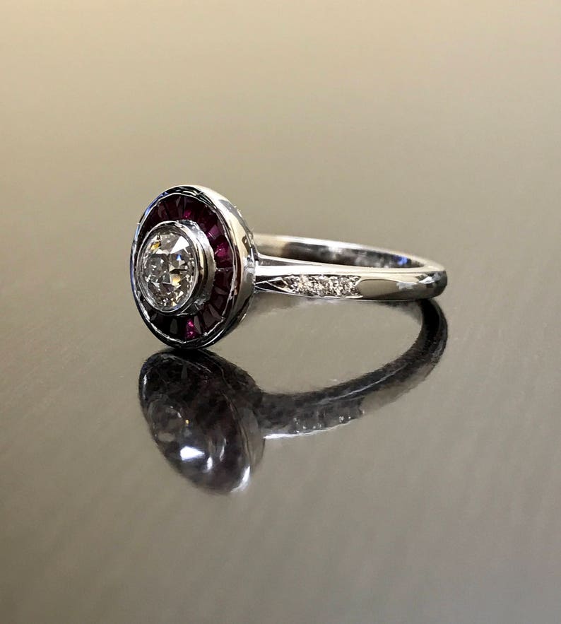 May include: A silver ring with a round, bezel-set diamond surrounded by a halo of red rubies. The band is set with smaller diamonds.