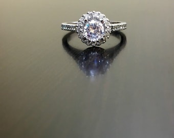 May include: A silver ring with a round, clear center stone surrounded by smaller stones. The ring has a halo setting and a band with small stones.