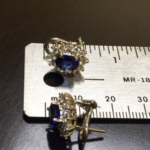 May include: A pair of silver earrings with a blue gemstone surrounded by smaller clear gemstones. The earrings have a clip-on closure.