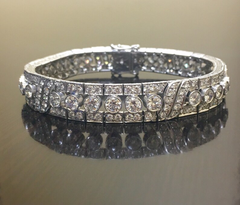 Puede incluir: Un brazalete de plata con una fila de diamantes redondos engastados en un dise&ntilde;o delicado e intrincado. El brazalete est&aacute; hecho de metal y tiene un acabado pulido.