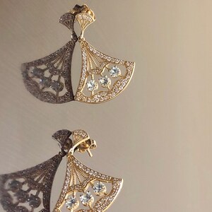 May include: A pair of gold earrings with a fan-shaped design and clear crystals. The earrings are delicate and elegant.