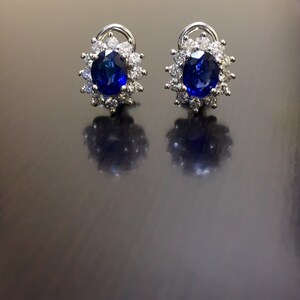 May include: A pair of silver earrings with a blue sapphire gemstone surrounded by a halo of smaller diamonds.