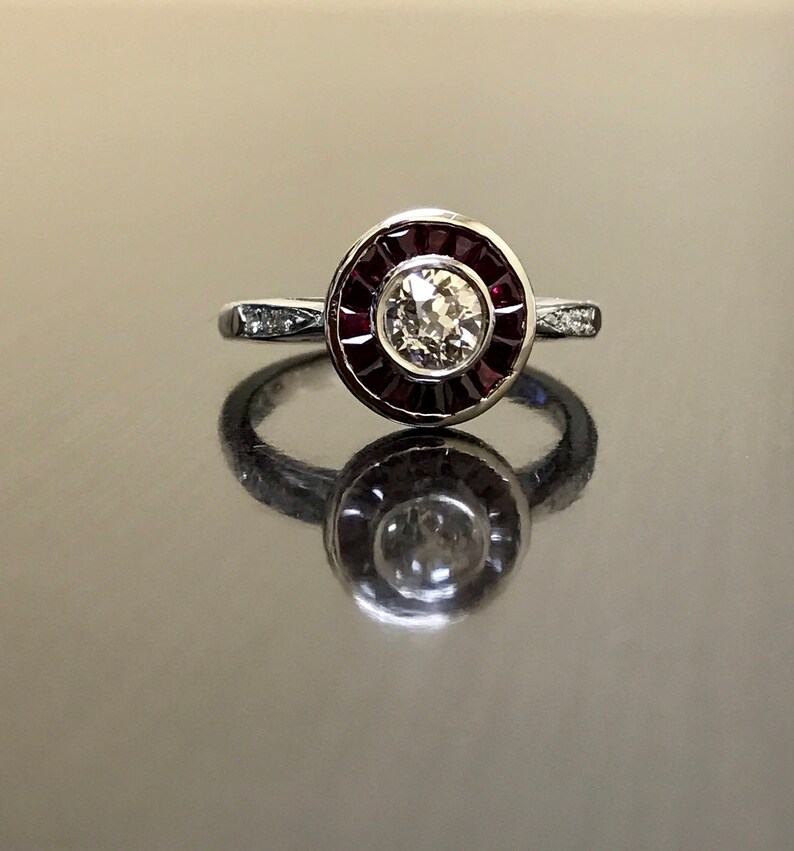 May include: A silver ring with a round, bezel-set diamond surrounded by a halo of red rubies.