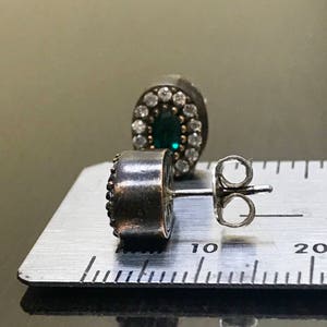 May include: A pair of silver earrings with a green gemstone surrounded by a halo of smaller white gemstones. The earrings are shown on a ruler with the measurement of 10 centimeters.