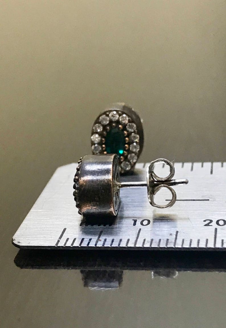 May include: A pair of silver earrings with a green gemstone surrounded by a halo of smaller white gemstones. The earrings are shown on a ruler with the measurement of 10 centimeters.