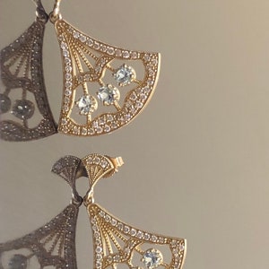 May include: A pair of gold earrings with a fan-shaped design. The earrings are adorned with clear gemstones and have a delicate, intricate design.