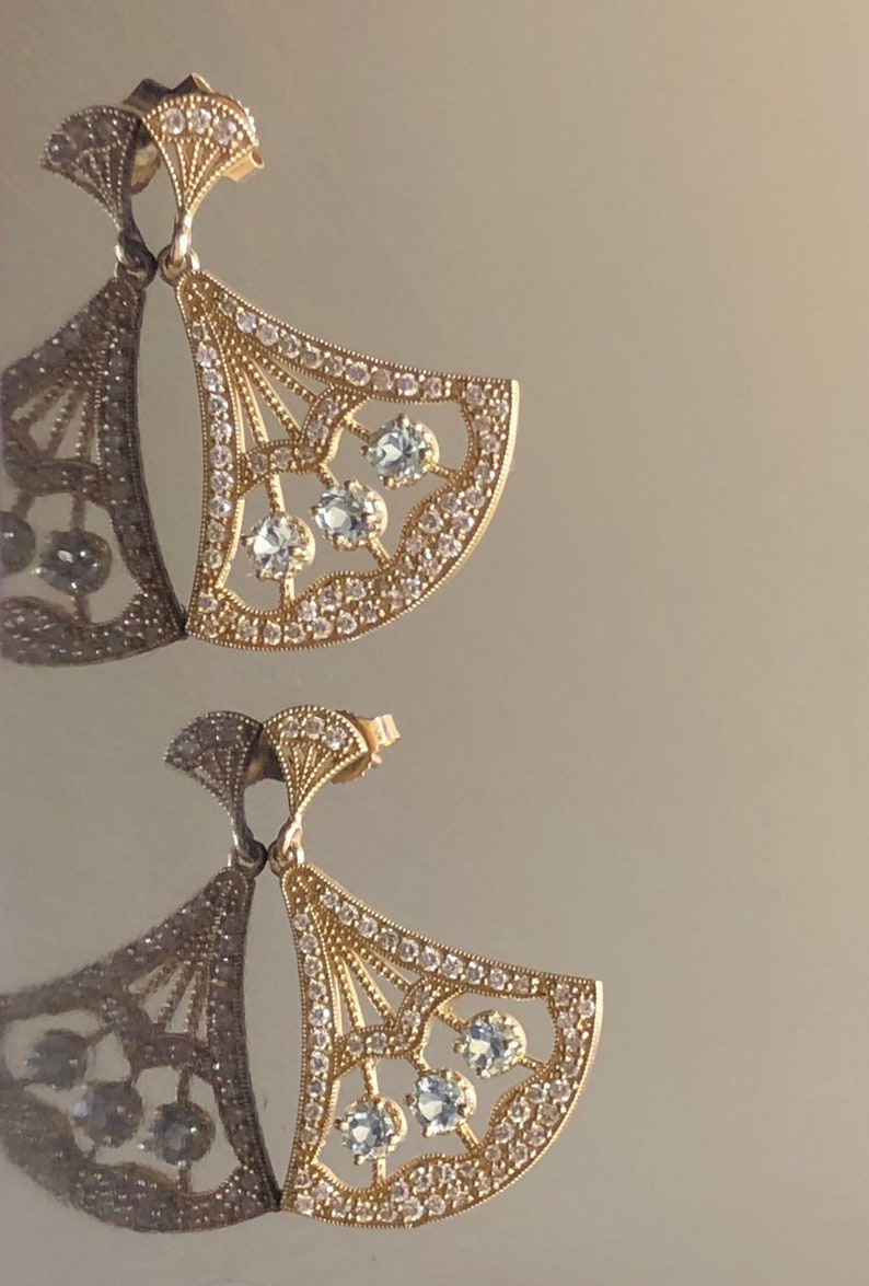 May include: A pair of gold earrings with a fan-shaped design. The earrings are adorned with clear gemstones and have a delicate, intricate design.