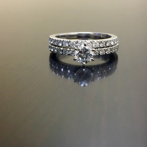 May include: A set of two diamond engagement rings. The top ring features a large, round, center diamond surrounded by smaller diamonds. The bottom ring is a simple band with a row of small diamonds.