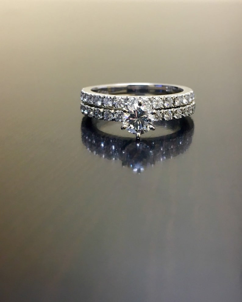 May include: A set of two diamond engagement rings. The top ring features a large, round, center diamond surrounded by smaller diamonds. The bottom ring is a simple band with a row of small diamonds.