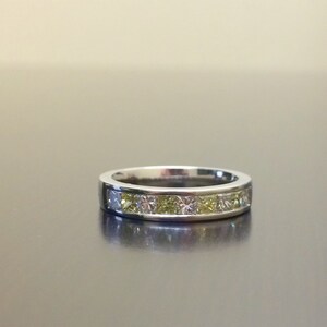 May include: A silver band ring with a row of small, square-shaped diamonds. The diamonds are a mix of clear and yellow.